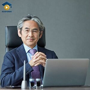 A professional man in a suit sitting at a desk with a laptop, visually defining the English vocabulary word "businessman" (a man who works in business) for the Read4More online glossary.